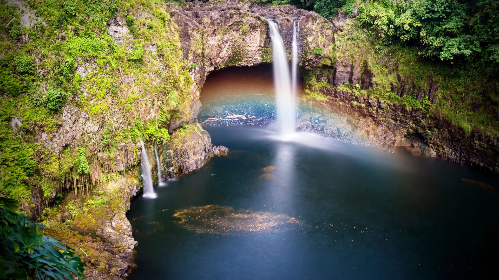 Waterfall cascading into a pool with a rainbow and lush greenery.